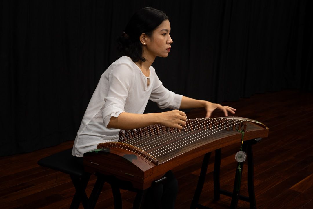 Léo on one knee reaching upward, a đàn tranh player behind him on a dark stage