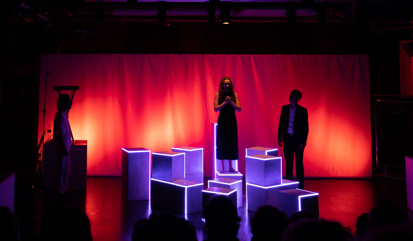 Wide shot of the stage under a red cyclorama, cubes lit from inside around the actors